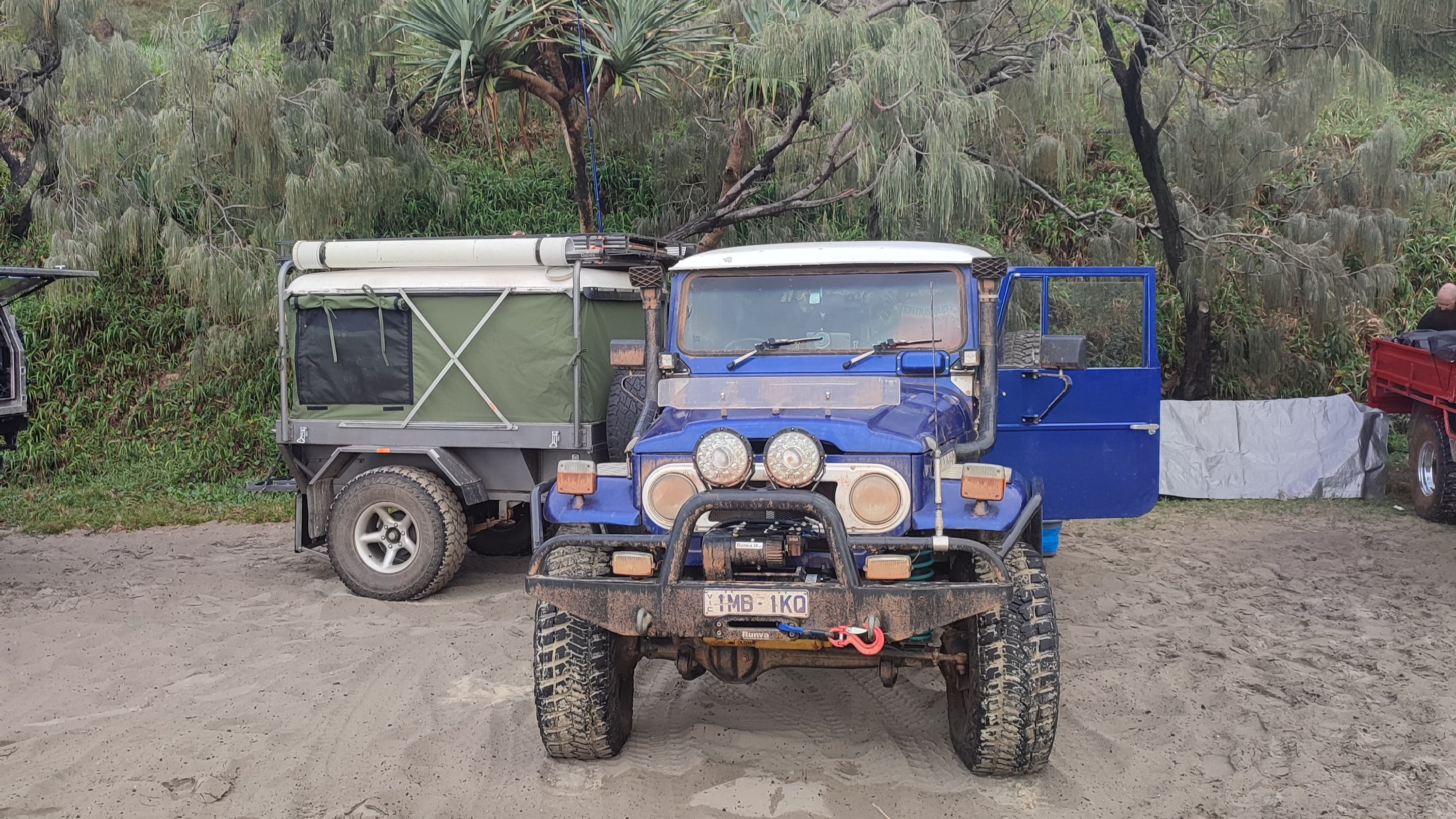 A blue 4x4 vehicle with a camper trailer on a sandy track near trees.
